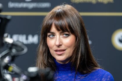 Dakota Johnson, de franja e blusa azul de renda, dá entrevista diante de câmeras e microfone
