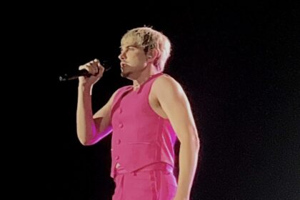 Jão canta ao microfone no palco, vestindo colete e calça cor-de-rosa