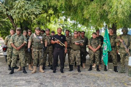 Grupo de veteranos do GVEB posa em fileira, em trajes militares, ao lado da bandeira do grupamento, sob árvores