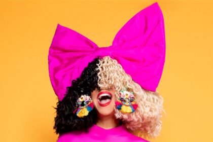 Sia com peruca metade preta e metade loira, laço rosa gigante na cabeça e vestido pink, em fundo laranja