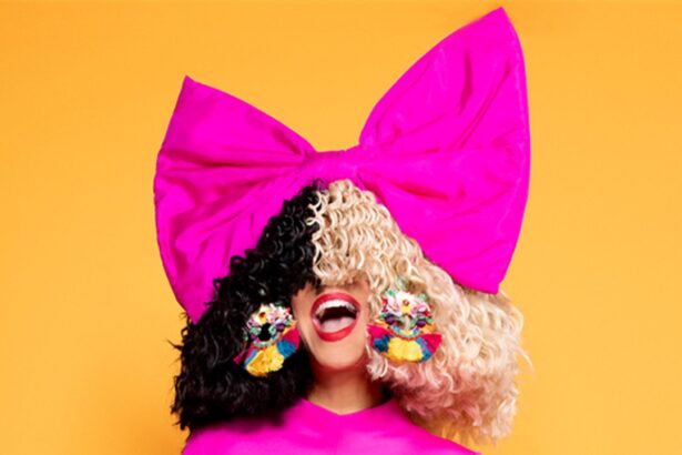 Sia com peruca metade preta e metade loira, laço rosa gigante na cabeça e vestido pink, em fundo laranja